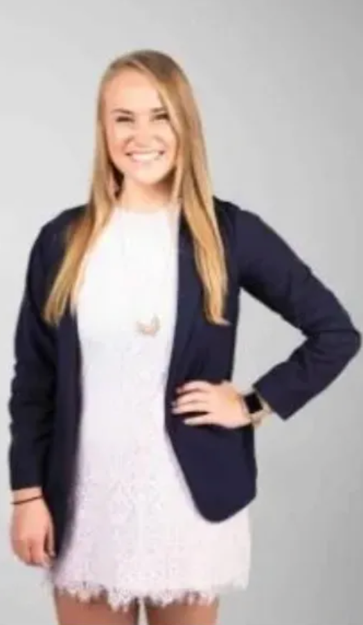 Woman with long blonde hair, smiling, in a white dress and navy blazer.