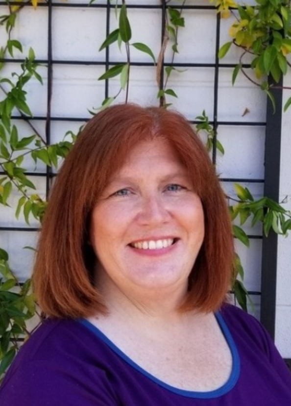 Woman with red hair smiles in front of a trellis with vines. She wears a purple shirt.