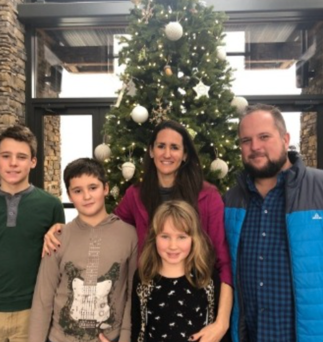 Family of five smiling in front of a decorated Christmas tree.