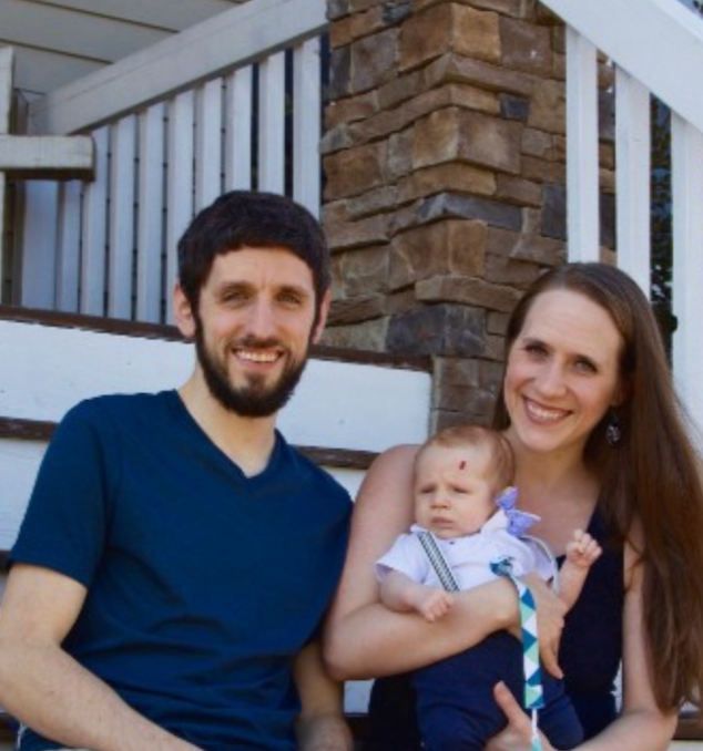 A smiling family of three on a porch. Man in blue, woman holding a baby.
