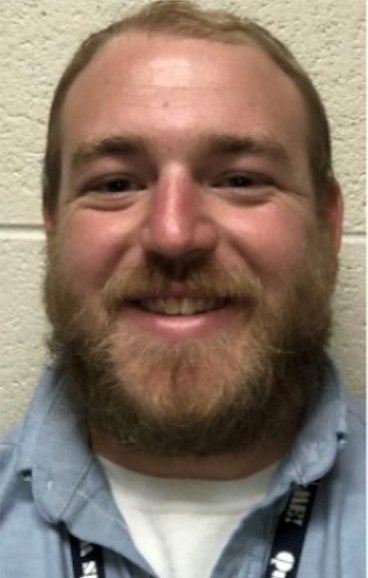Man with a beard smiles at the camera, wearing a blue collared shirt. Light brick wall background.