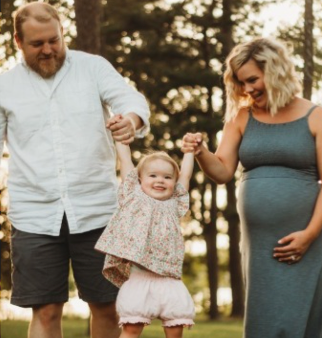 Family walking outdoors, holding hands. Pregnant mother and father smiling, lifting up a toddler.