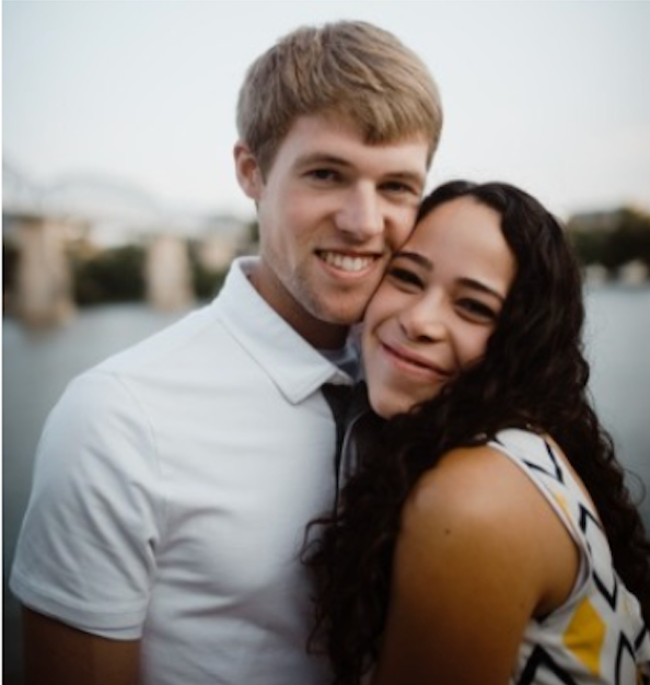 Smiling couple embraces by water, man in white shirt, woman in patterned top.