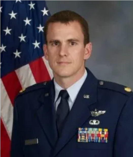 US Air Force officer in uniform, standing in front of an American flag.