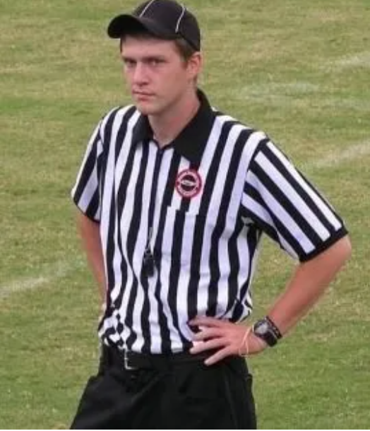 Referee on a green field, wearing black and white striped shirt, black cap, hands on hips, serious expression.