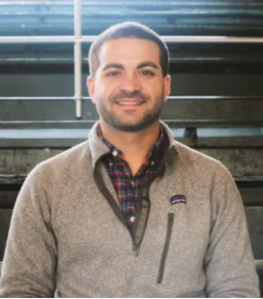 Man in grey sweater, smiling, wearing plaid shirt, in a setting with industrial-style background.