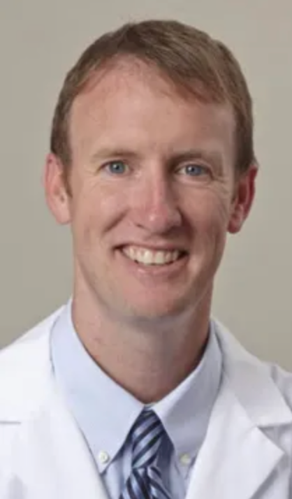Smiling fair-skinned man with reddish hair, wearing a white lab coat, blue striped tie, and blue collared shirt.