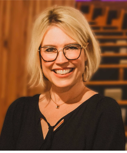 Blonde woman with glasses smiles in a studio, wearing a black top and earrings.