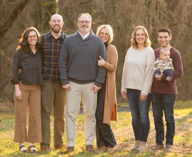 Family poses outdoors, sunny, includes baby held by young man.