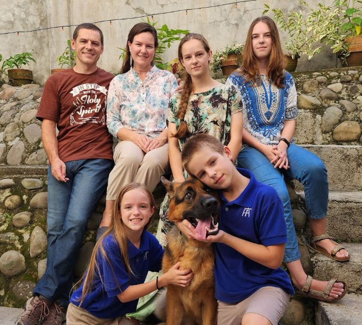 Family of six with a German Shepherd dog on stone steps; smiling, outdoors.
