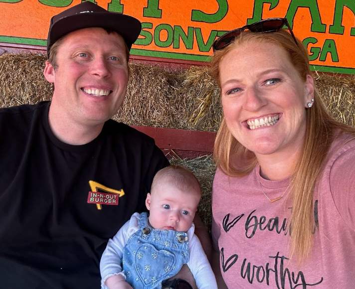 Family of three smiling at the camera in front of hay bales; baby in denim overalls.