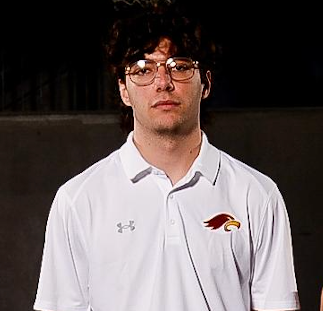Man with glasses wearing a white polo shirt with a logo; dark background.