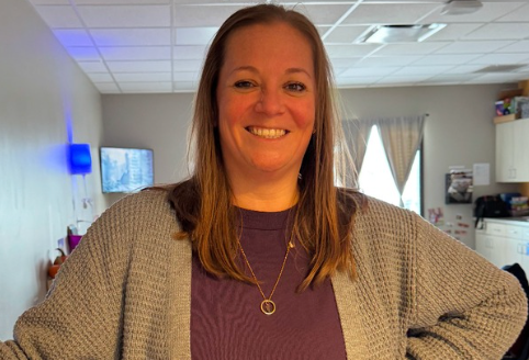 Woman with long brown hair, smiling, wearing a purple shirt and gray cardigan in a room.