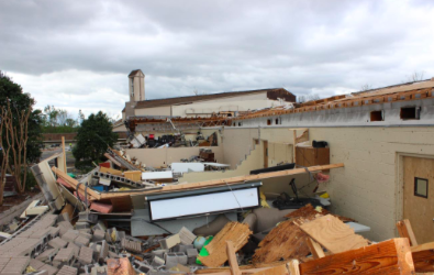 Building partially collapsed; debris piles up. Exterior view of damaged structure under cloudy skies.