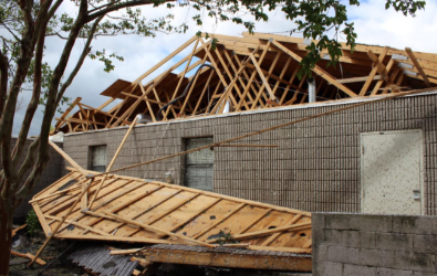 Damaged building after a storm; roof torn off, exposing rafters and wood frame. Brick exterior, cloudy sky.