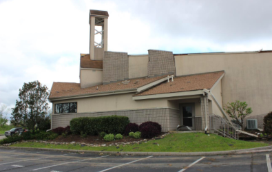 Church building with beige walls, brown roof, a bell tower, and landscaped lawn.