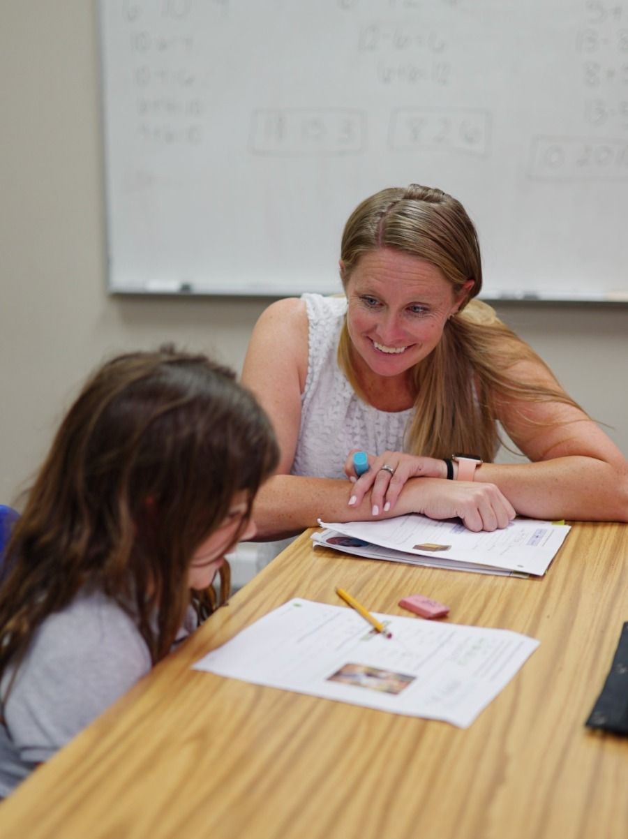 A woman smiles at a young girl, helping her with schoolwork at a desk in a classroom.