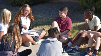 A group of diverse students sits in a circle outdoors, studying with books and smiling.