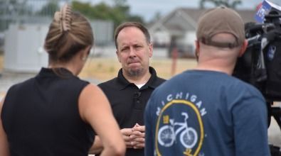 A man in black shirt looks down while being interviewed. Two people stand in front, a woman and a man with Michigan shirt.