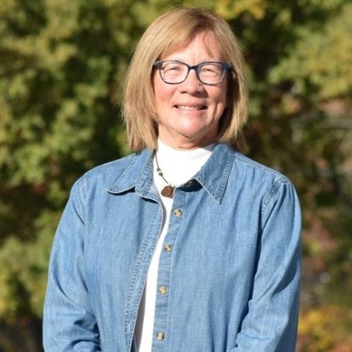 Woman with glasses, blonde hair, denim shirt, white turtleneck, smiles in front of a tree.