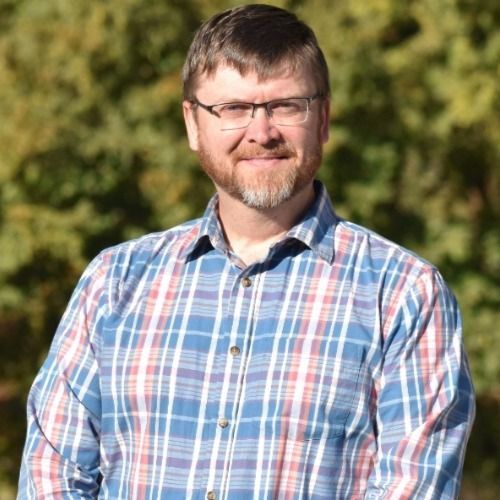 Man with glasses and beard smiles outdoors, wearing a plaid shirt.