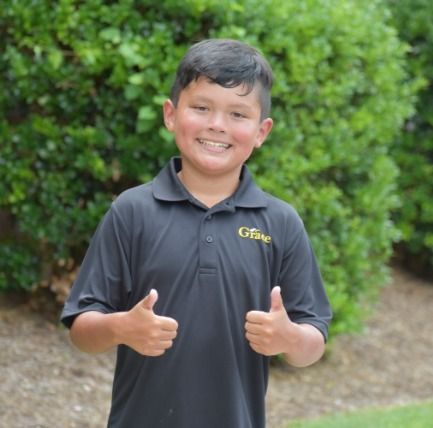 Boy in black shirt with thumbs up, smiling outside near green bushes.