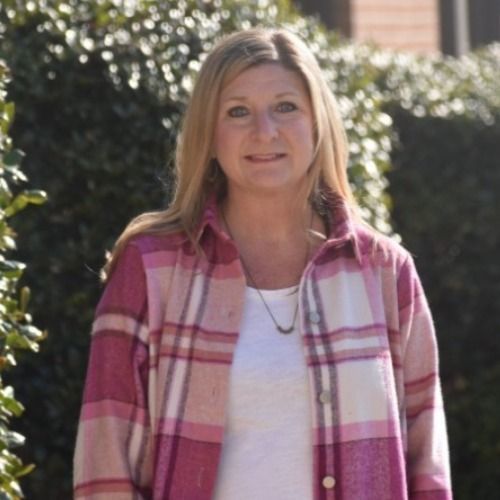 Woman with blonde hair, wearing a pink plaid shirt over a white top, smiles outside.