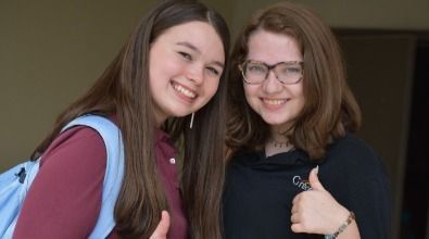 Two smiling girls giving thumbs up. One with backpack, one wearing glasses.