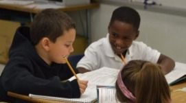 Two boys smiling, working together at a desk with another person; writing and looking at a notebook.