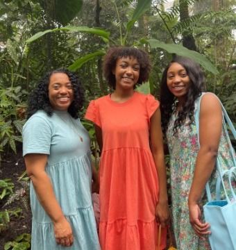 Three Black women pose in lush greenery. One in blue, one in orange, and one in a floral dress smile.