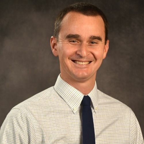 Man in white checkered shirt and dark blue tie smiles at the camera.