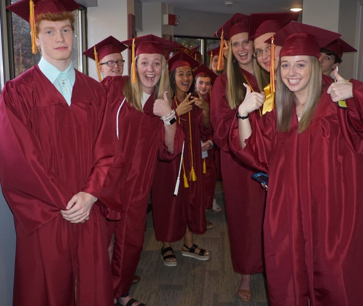 Graduating students in maroon gowns and caps, smiling and posing for a photo in a hallway.