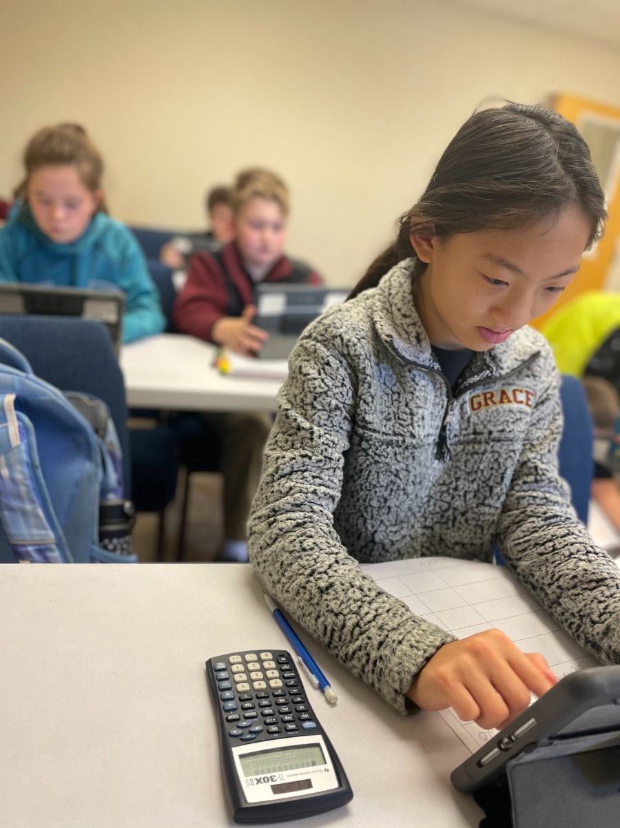 Students in a classroom working on assignments. Girl in sweater at the front with a calculator, tablet, and paper.