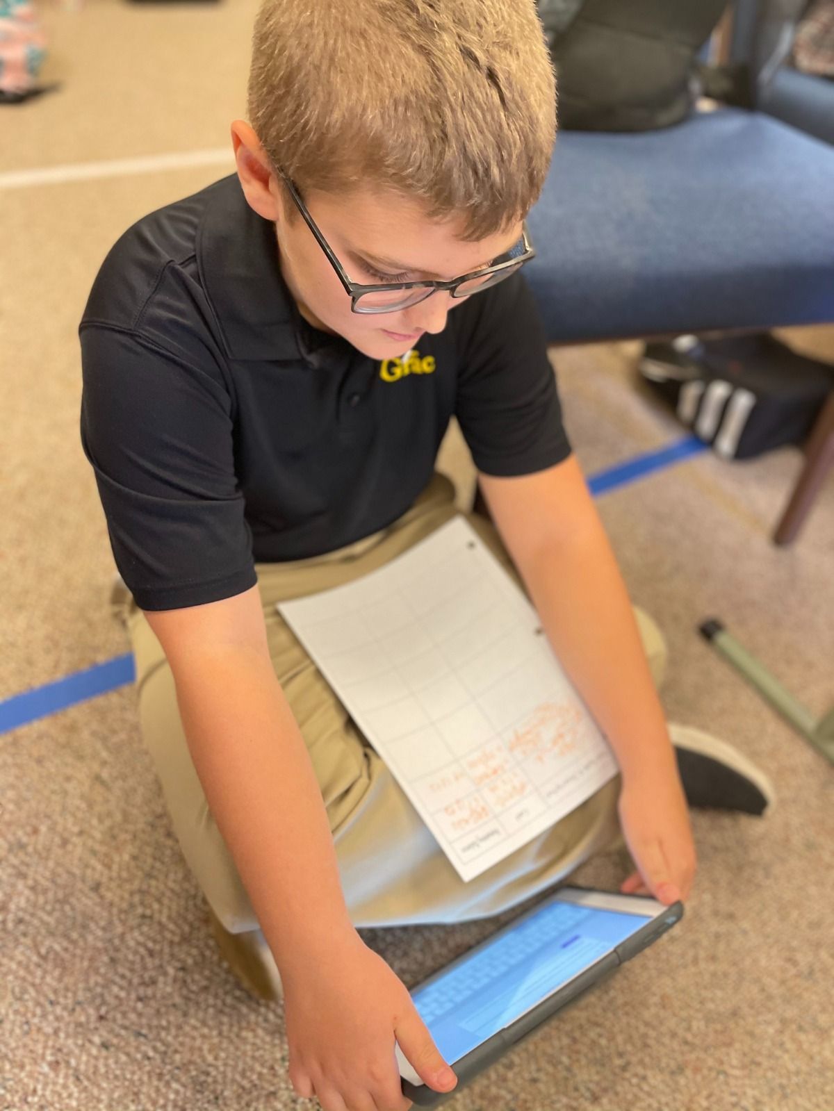 Boy in glasses sits cross-legged on the floor looking at a tablet, paper on his lap.