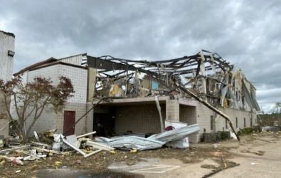 Building destroyed by a storm; roof collapsed, debris scattered in front. Overcast sky.
