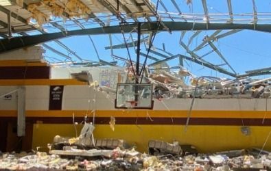 Basketball hoop in a damaged gym, roof collapsed, debris scattered. Yellow, brown, white walls.