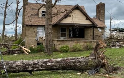 House damaged by a storm, tree fallen in front. Brick and beige house with missing windows.