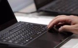 Hand typing on a black laptop keyboard, another laptop in the background.