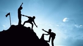 Silhouetted figures helping each other climb a mountain to reach the flag on top.