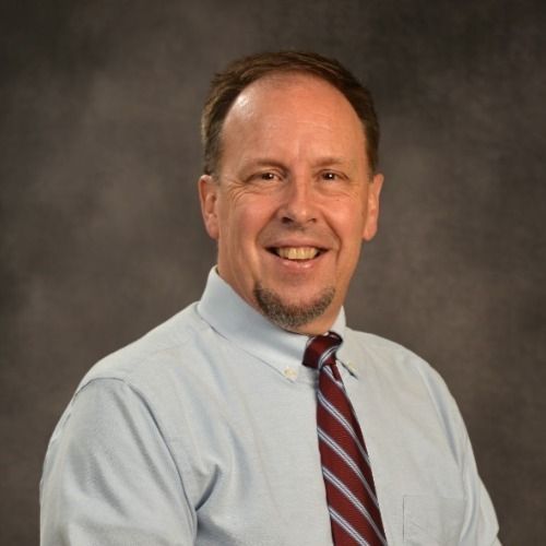 Smiling man with goatee, wearing a blue button-down shirt and patterned tie.