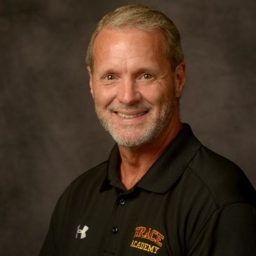 Man with gray hair and beard smiling, wearing a black Grace Academy polo shirt.