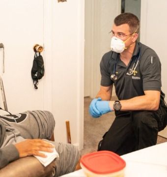 Paramedic kneels, examines patient's leg, wearing mask and gloves in a room.