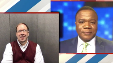 A split screen shows two men. One man smiles, wearing glasses, a burgundy vest, and a white shirt. The other man, in a suit, looks serious.