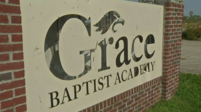 Sign for Grace Baptist Academy, beige with black text and eagle logo, set in front of brick.