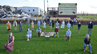 Graduating seniors pose for photo on field, some wearing masks, in front of damaged school.