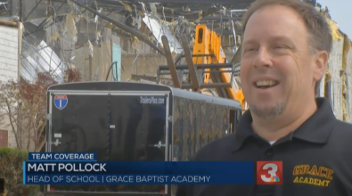 Matt Pollock, Head of School, smiling in front of damaged building. Grace Baptist Academy sign, trailer, and construction equipment.