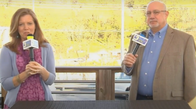 Two people, a woman and a man, holding microphones, standing outside, possibly for a news broadcast.