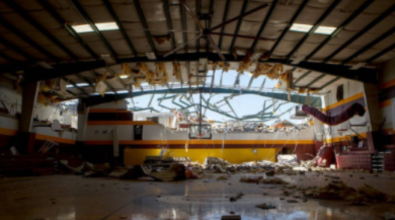 A gymnasium with a collapsed roof and debris scattered on the floor after a disaster.