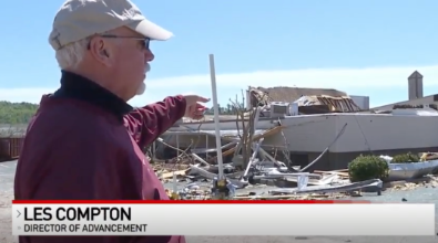 Man in cap points at tornado-damaged building.