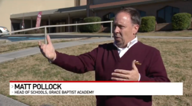 Matt Pollock, Head of Schools at Grace Baptist Academy, gestures outside a school building, wearing a maroon sweater.